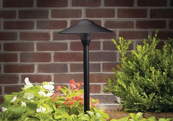 Black outdoor path light illuminating garden flowers against a brick wall.