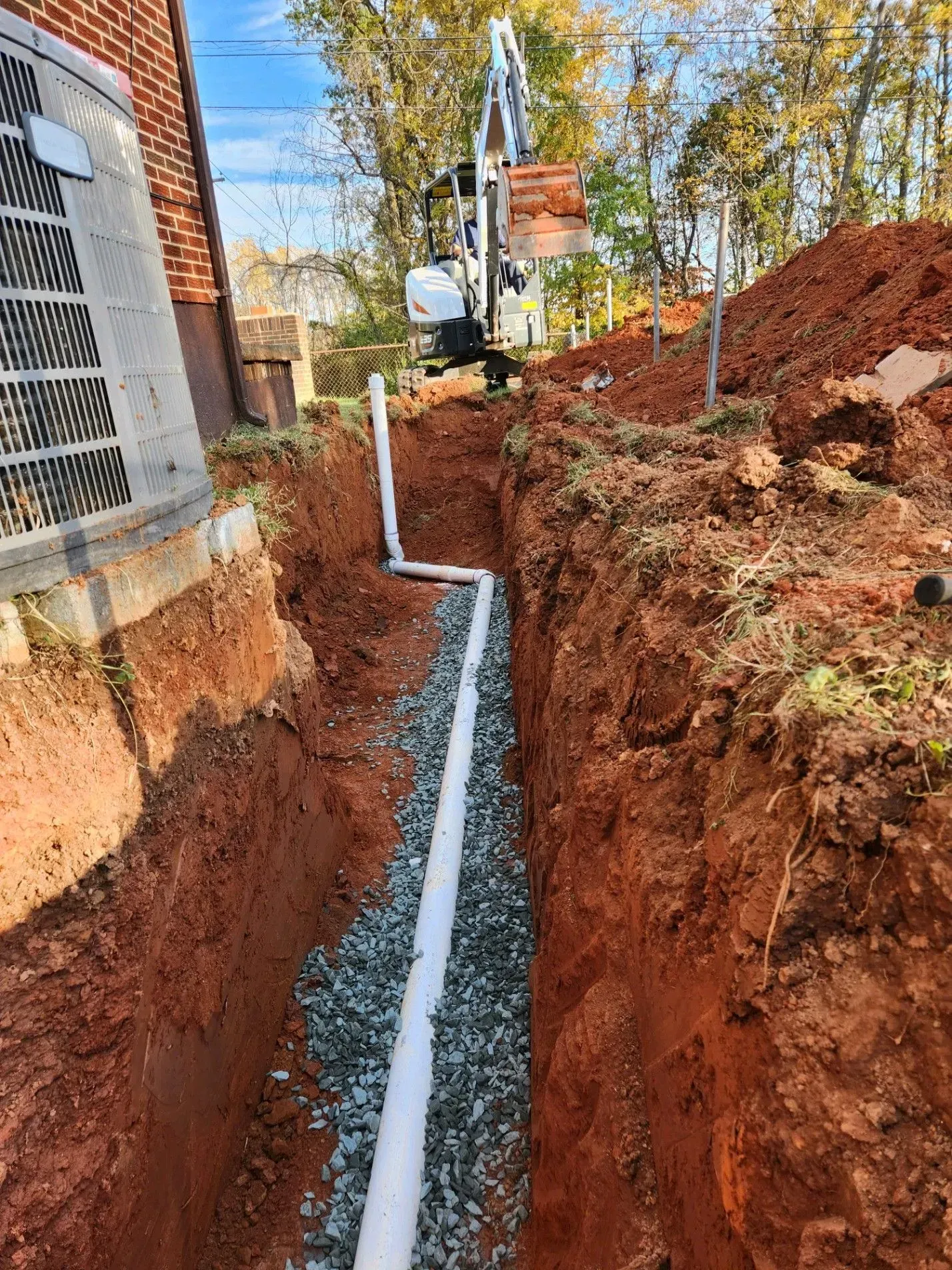 Trench with white pipes and gravel, near a building. An excavator is in the background.
