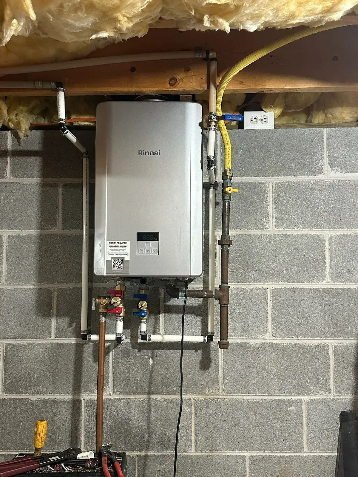 A wall-mounted, silver tankless water heater with plumbing against a cinder block wall.
