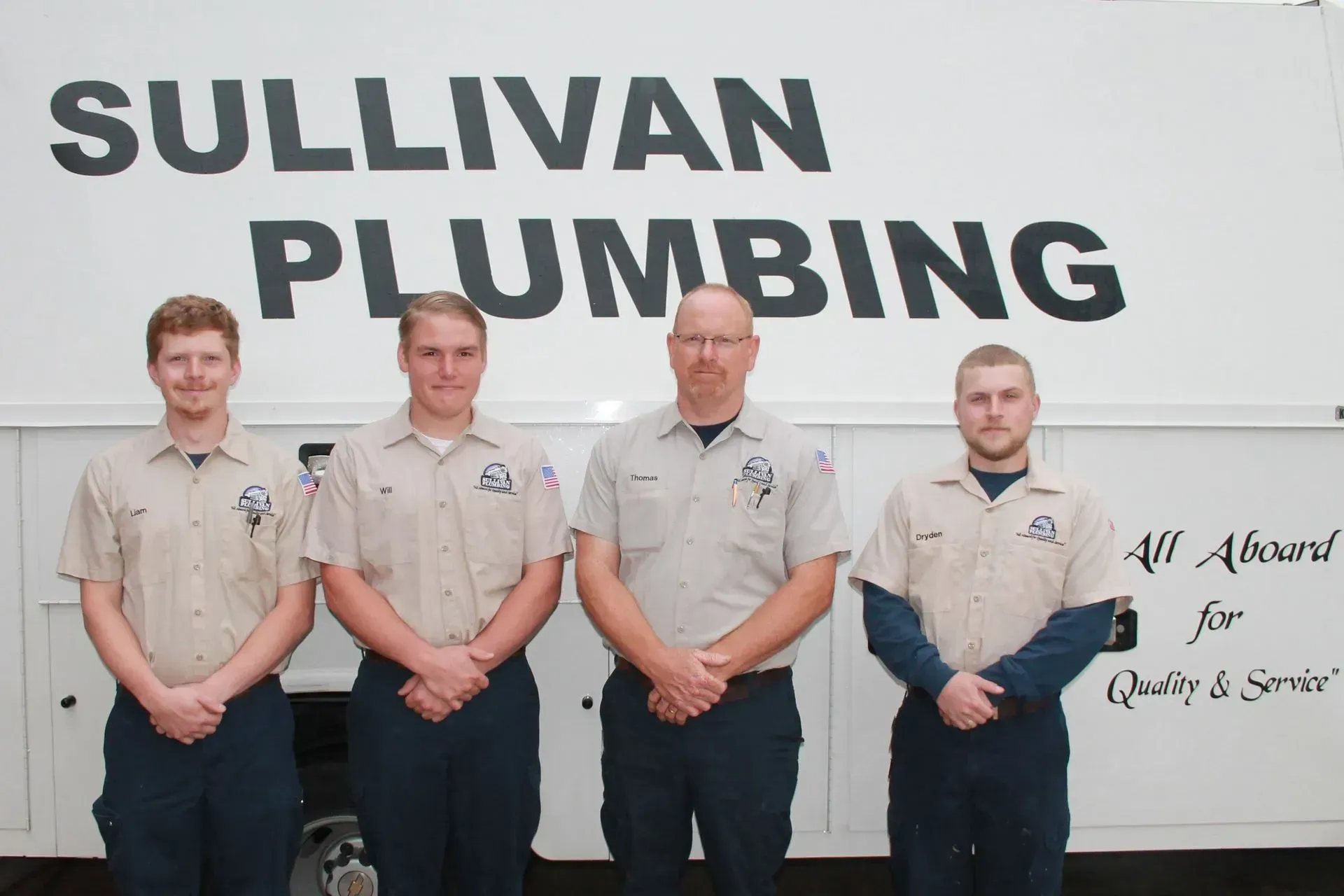 Four men in tan shirts and navy pants stand in front of a white truck with 