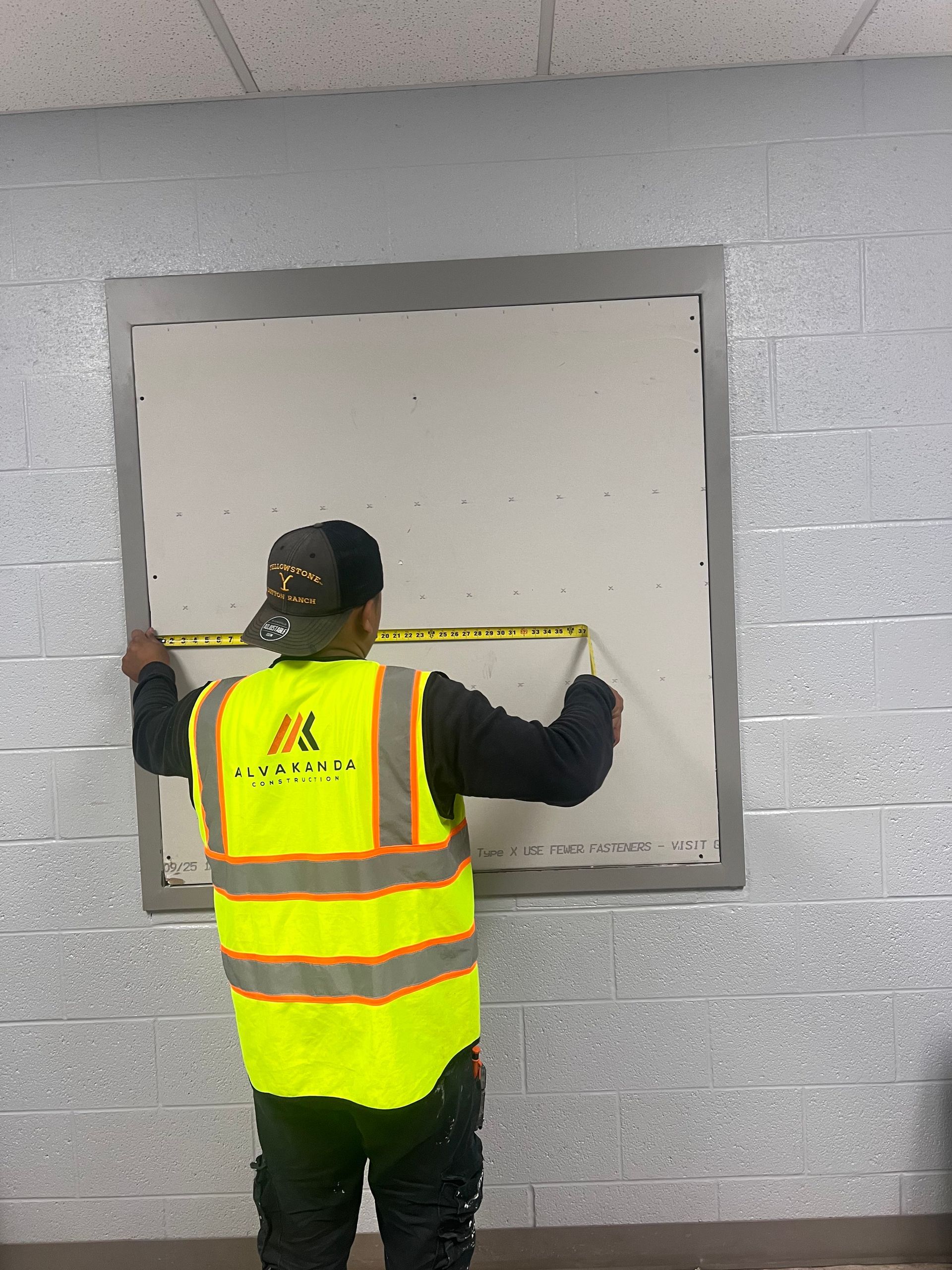 A man in a yellow vest is measuring a wall with a tape measure.