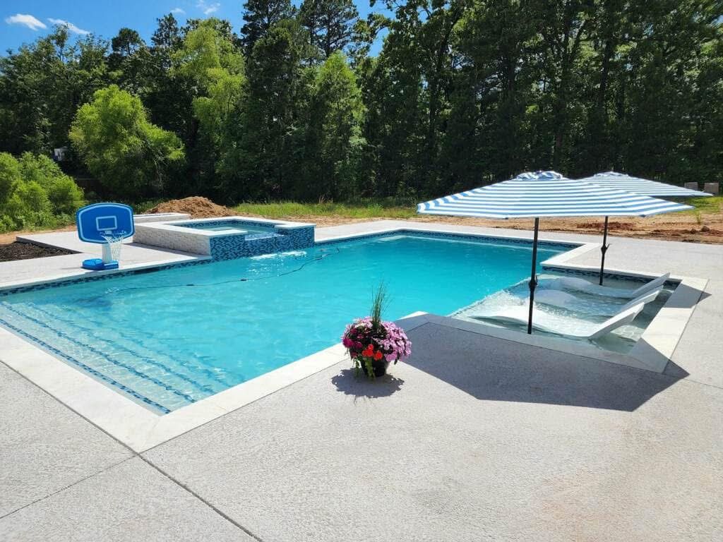 A large swimming pool is surrounded by trees and a patio with umbrellas.