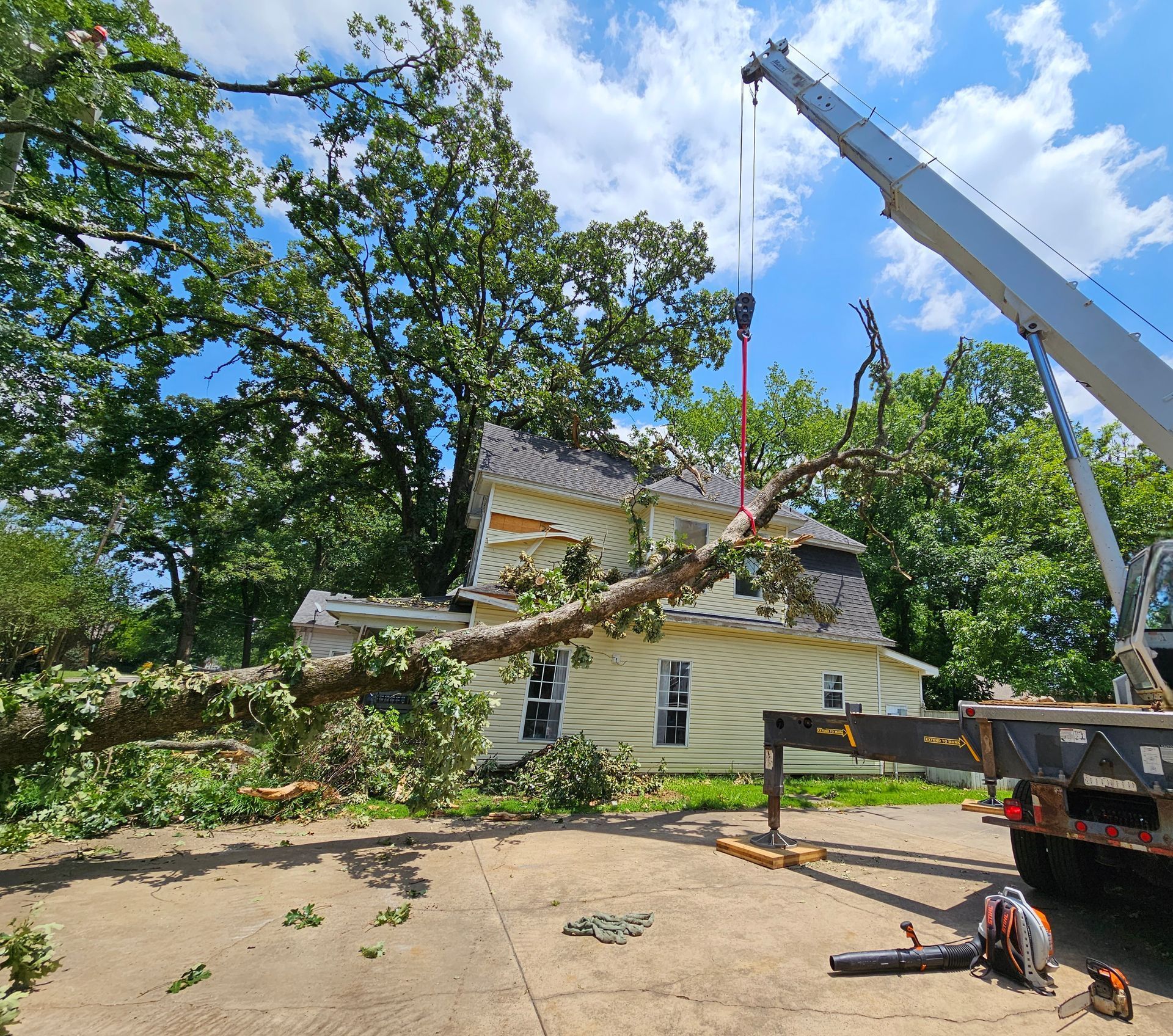 A crane lifts a large tree branch away from the roof of a pale yellow two-story house after storm damage.