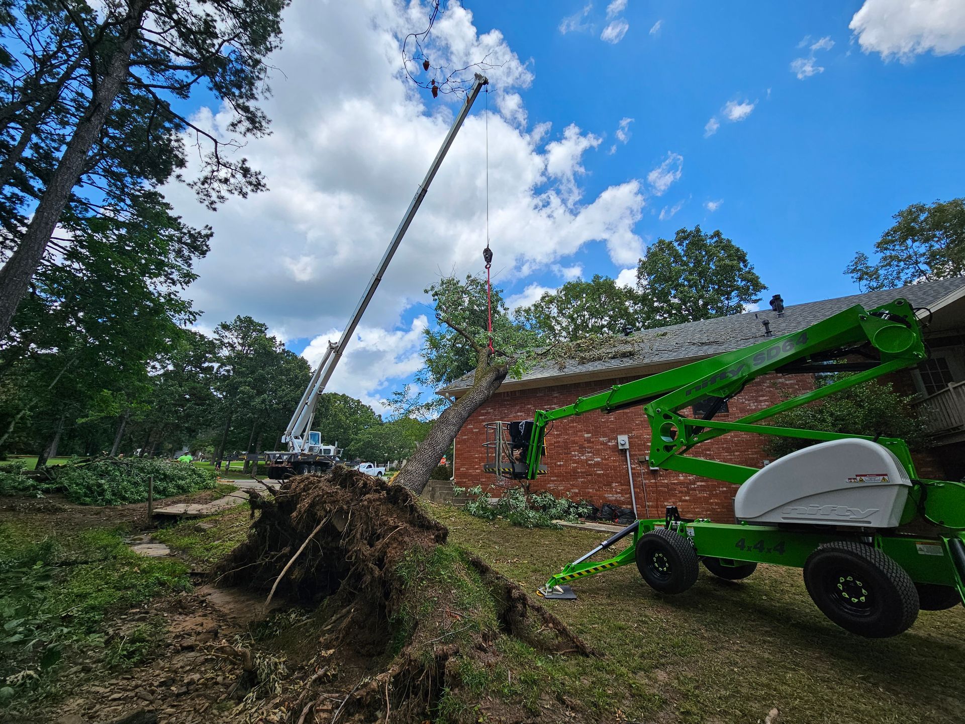 A crane removes a large fallen tree from the roof of a brick house, while a green aerial lift stands nearby on the lawn.