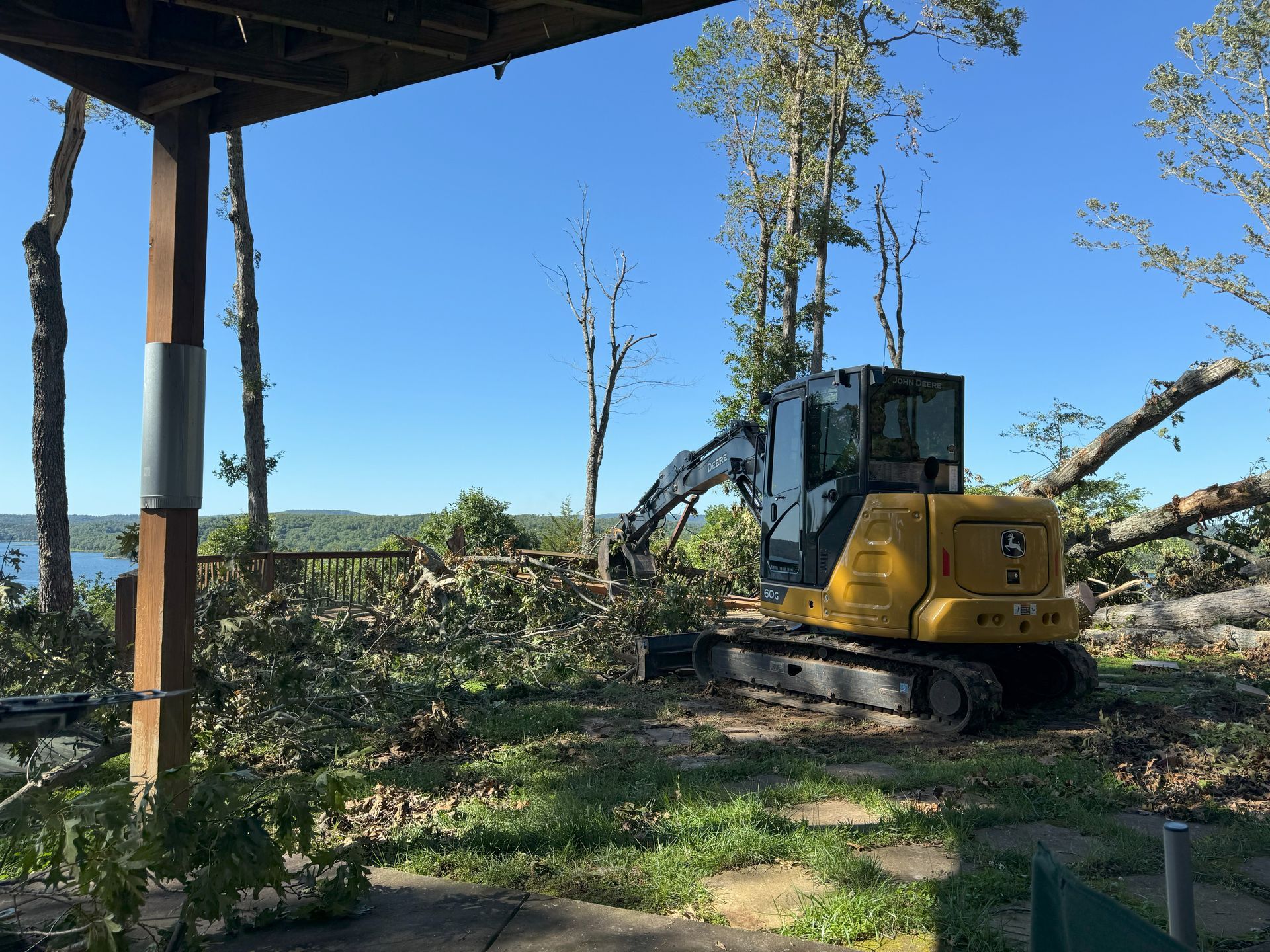 A yellow John Deere excavator works on clearing fallen trees and debris in a wooded area overlooking a lake.