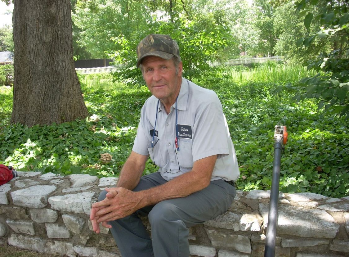 A person in a light-colored uniform and camo cap sitting on a stone wall in a garden setting, holding a long-handled tool.
