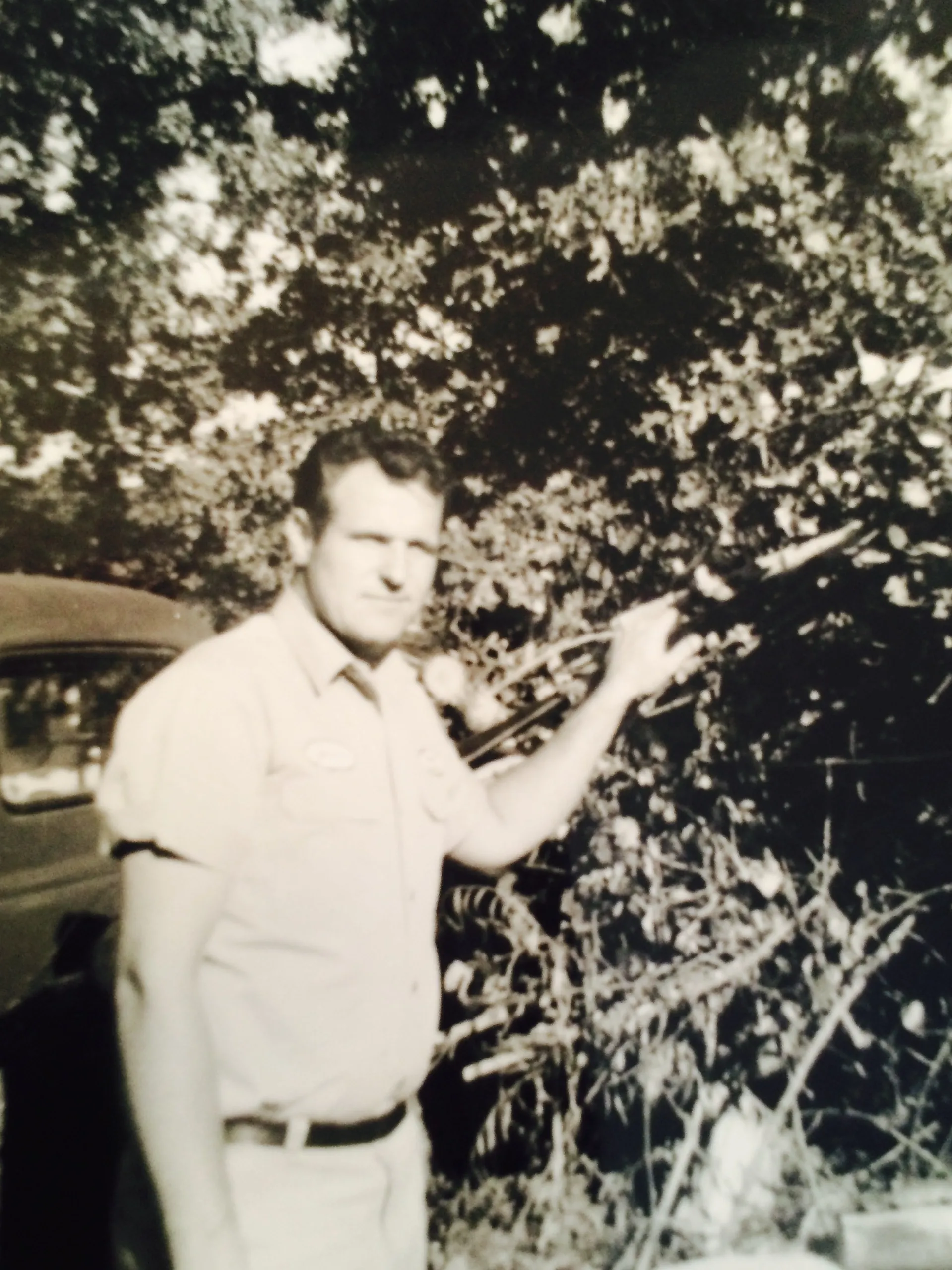 Man in light-colored shirt points to bushes, stands near a vehicle, outdoors in the shade.