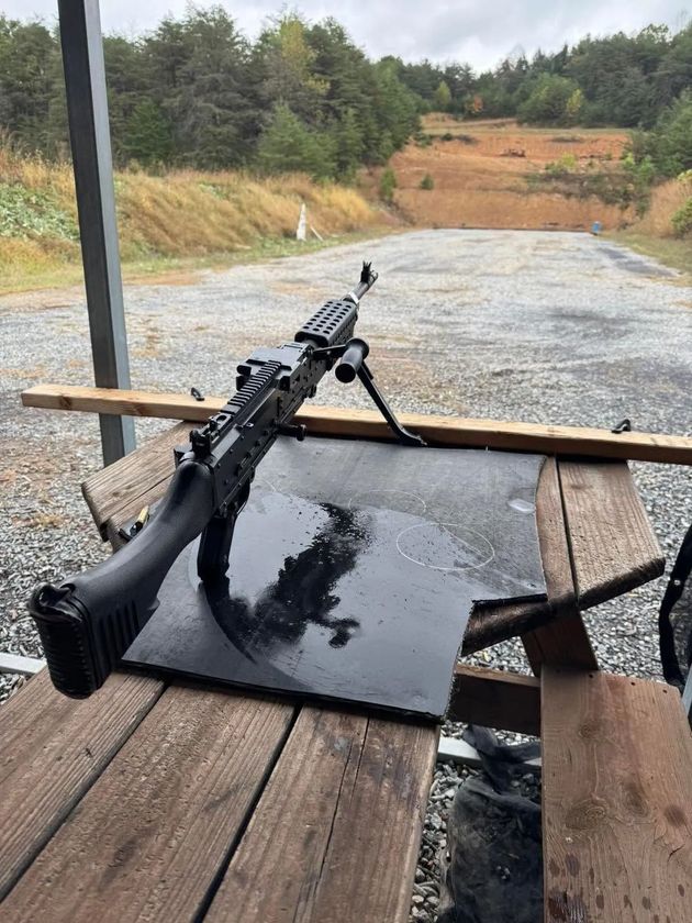 Rifle on a shooting bench with a glossy black plate, overlooking a gravel range and wooded hillside.