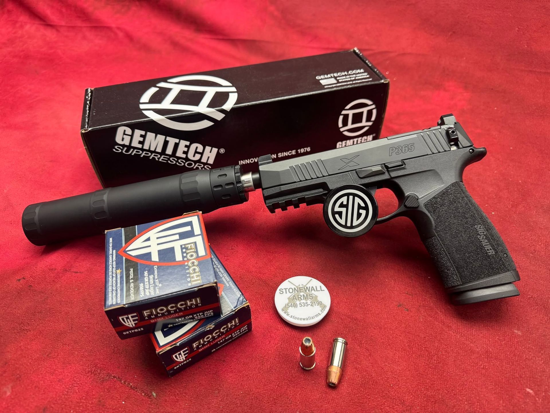 Suppressed black handgun with accessories on red cloth, boxed ammo, and a white coin-like token