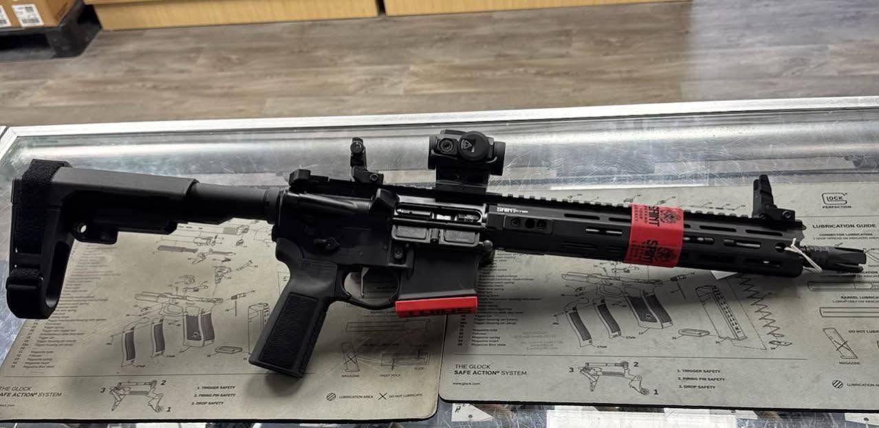 Black rifle with scope and red tape resting on a workbench next to a red tool or magazine.