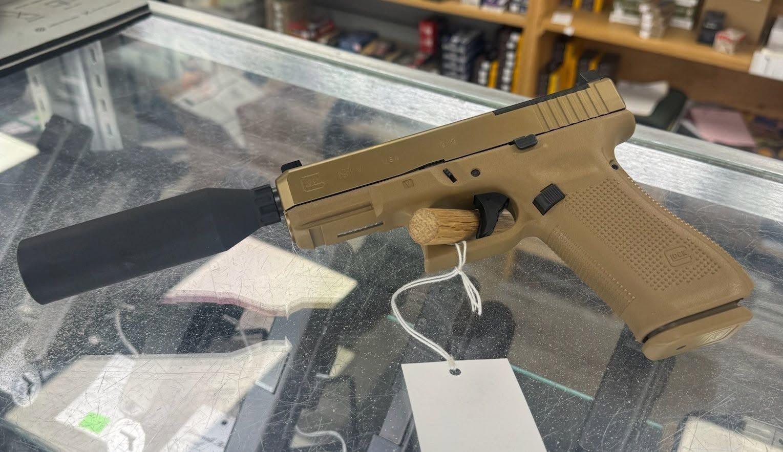 Tan handgun with a black suppressor displayed on a glass counter in a store.