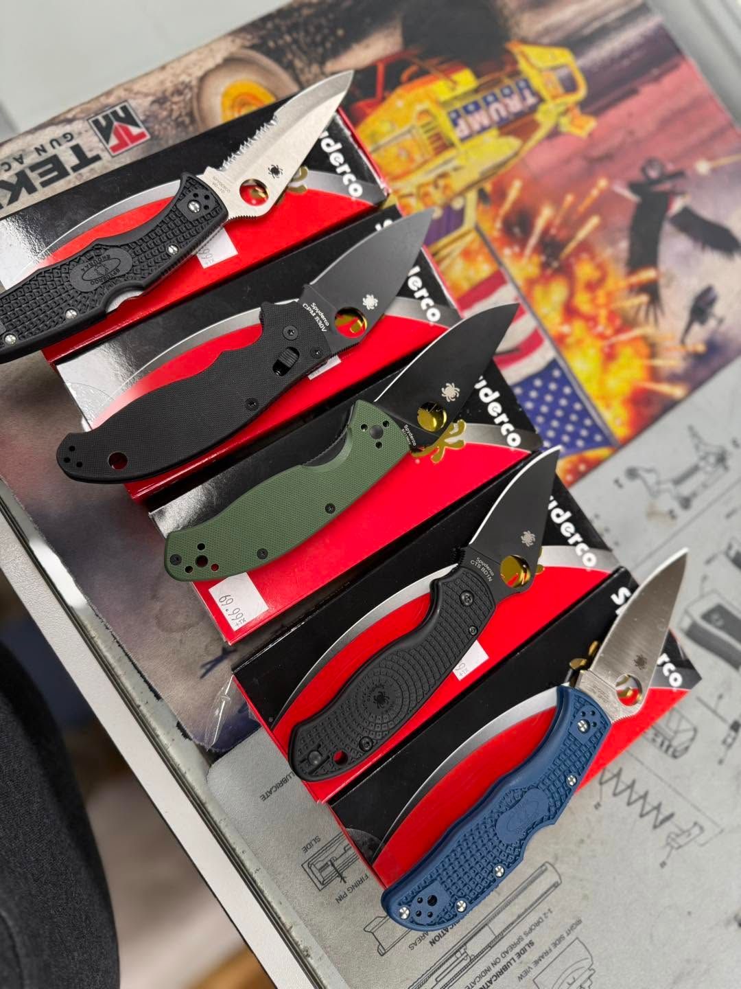 Six folding pocket knives arranged diagonally on a display, with black, red, green, silver, and blue handles.