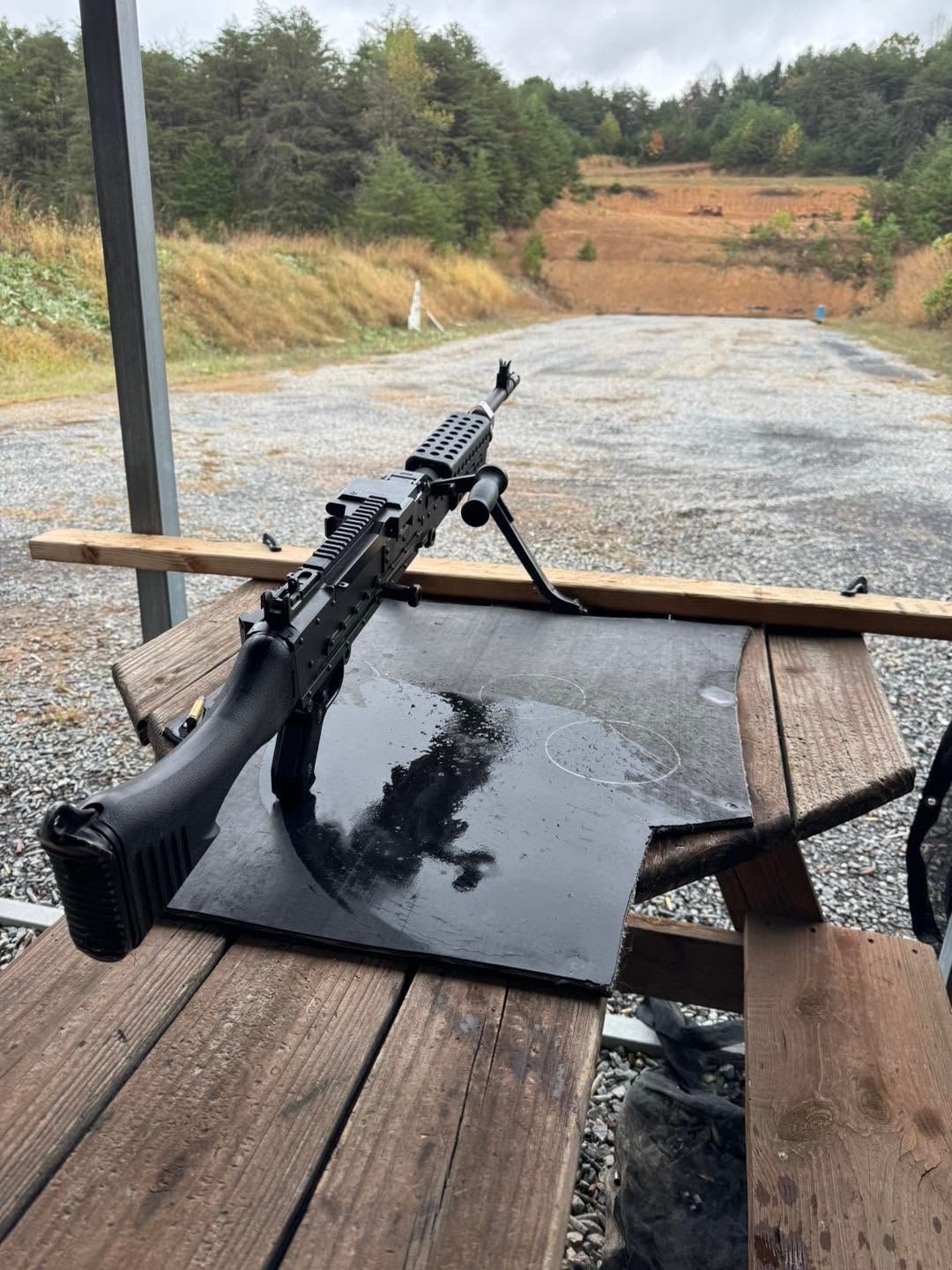 Black rifle on a wet wooden bench at an outdoor shooting range, with target berms in the background