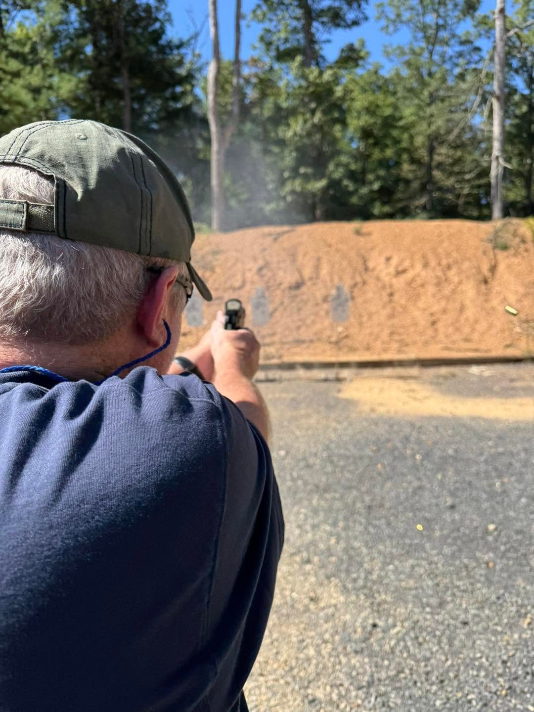Person aiming a handgun at an outdoor shooting range with a dirt backstop and trees.