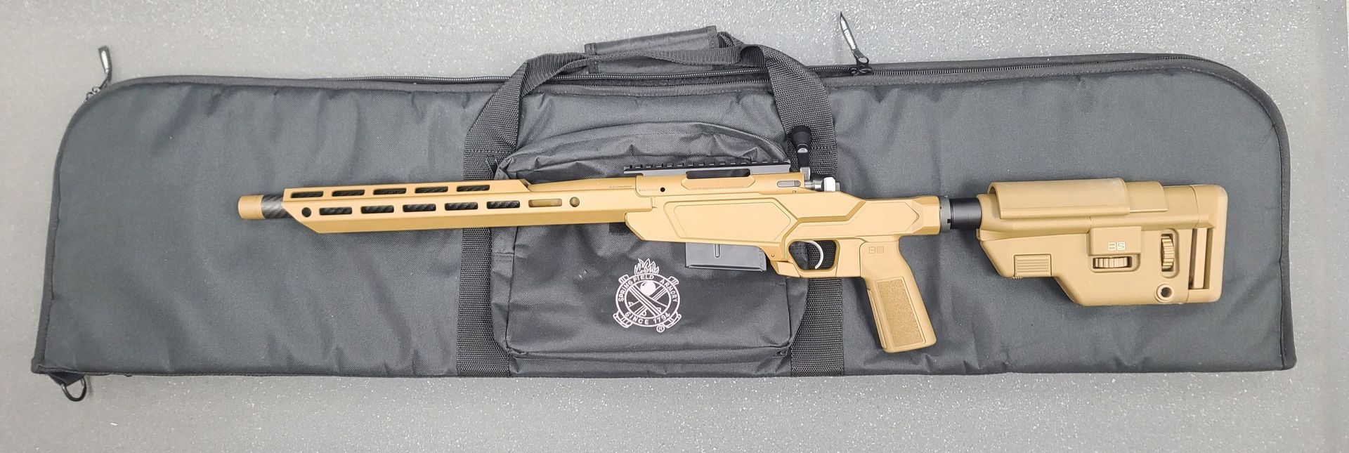 Tan rifle with scope and folded stock lying on a black soft case