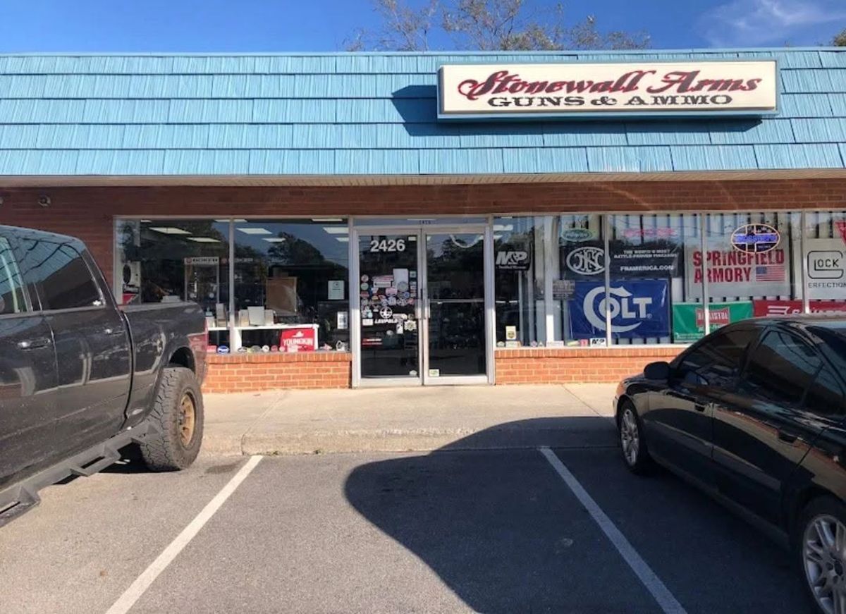 Strip mall storefront with blue awning, parked cars, and a glass entrance under a sign reading “International Arms.”