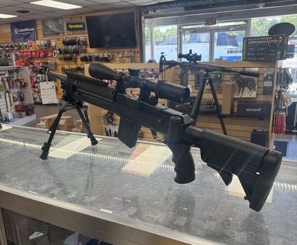 Two scoped rifles on a display table inside a gun shop, with ammo and accessories in the background