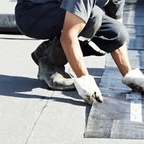Worker kneeling on a rooftop, smoothing black roofing material along a white edge