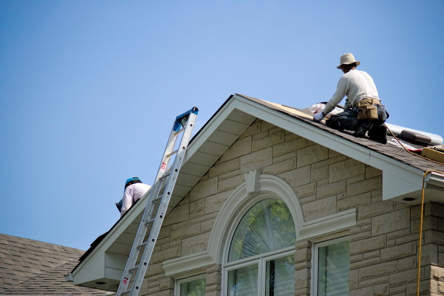Two roofers repairing a house roof, one on a ladder and one kneeling near the peak
