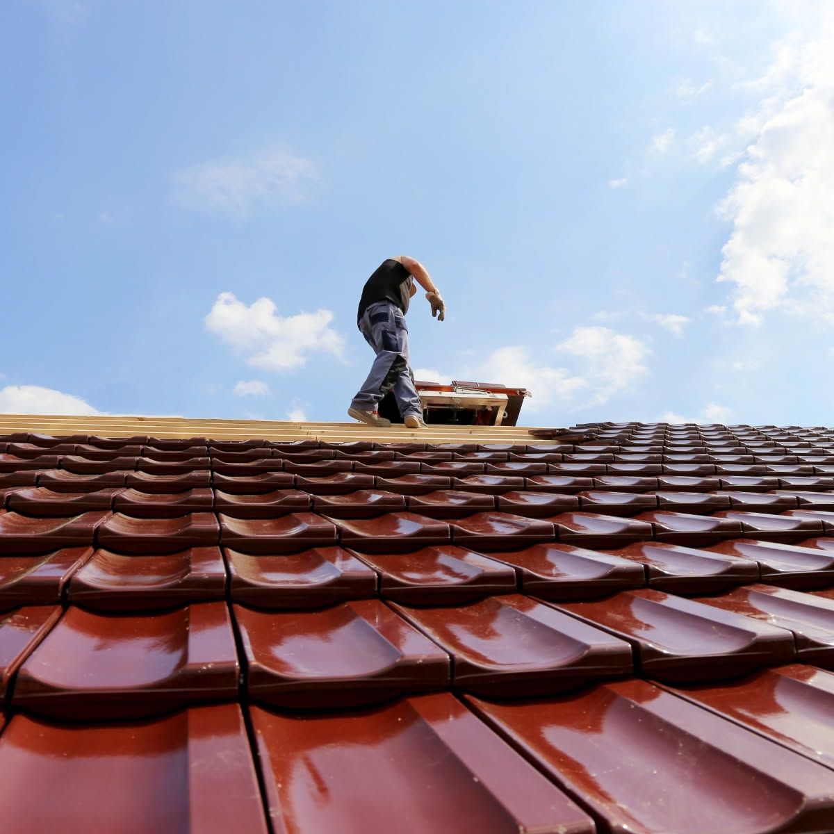 Worker on a red tiled roof beside a ladder, under a blue sky with clouds