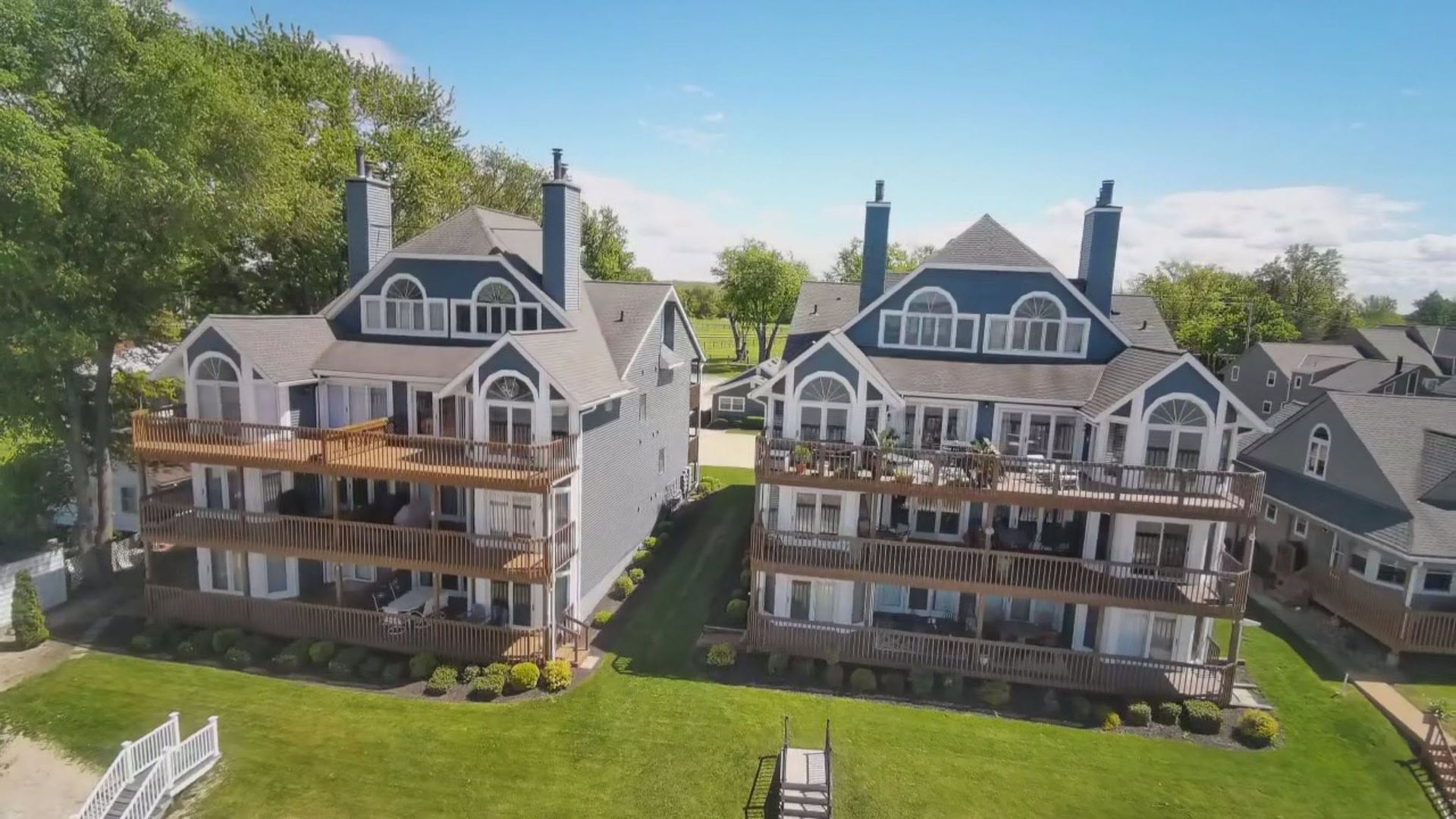 Two large gray houses with balconies beside a green lawn under a blue sky.