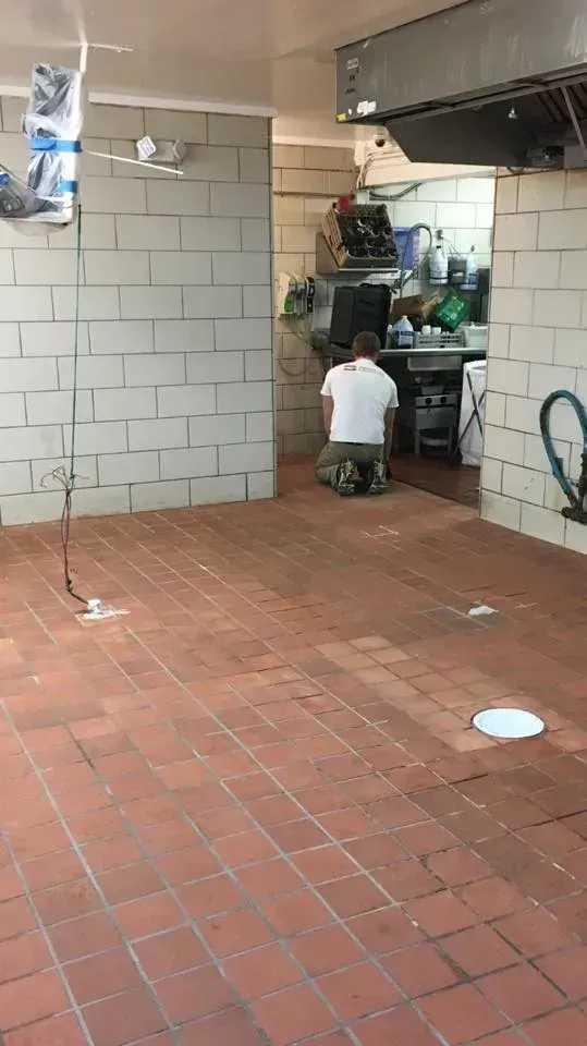 Person kneeling in a brick-floored restaurant hallway, looking into the kitchen. White brick walls.