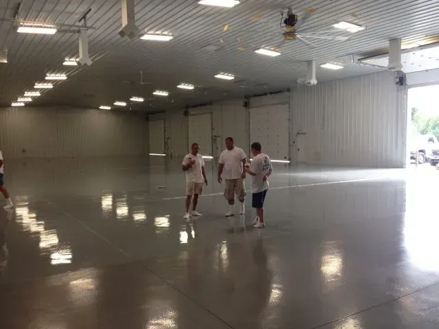 Three men in white shirts and shorts stand in a large, empty warehouse with a shiny gray floor.