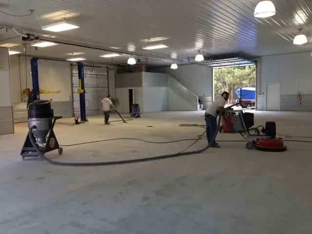 Workers cleaning a large, empty garage. One vacuums, another uses a floor polisher, and a third sweeps.