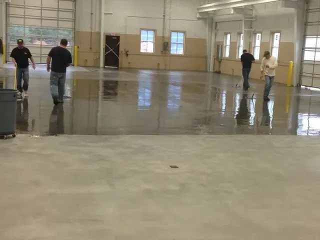 Four people walking on a shiny, freshly sealed concrete floor in a large, empty industrial building.