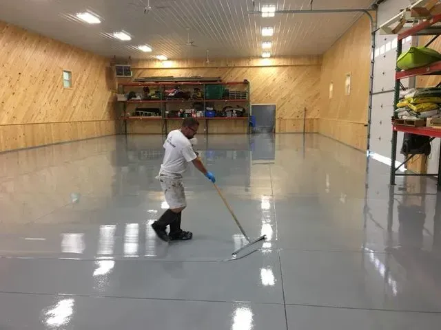 Man applying gray epoxy coating to a large interior floor with a long-handled squeegee.
