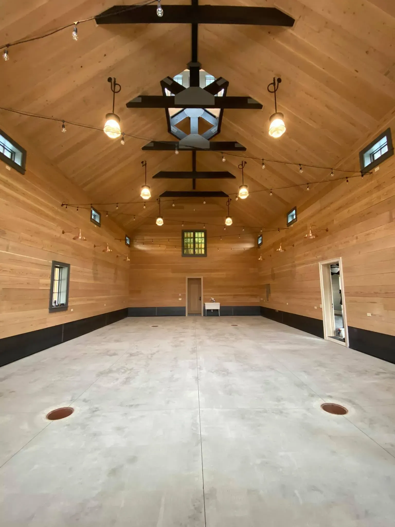 Interior of a large, empty wooden barn with string lights, concrete floor, and small windows.