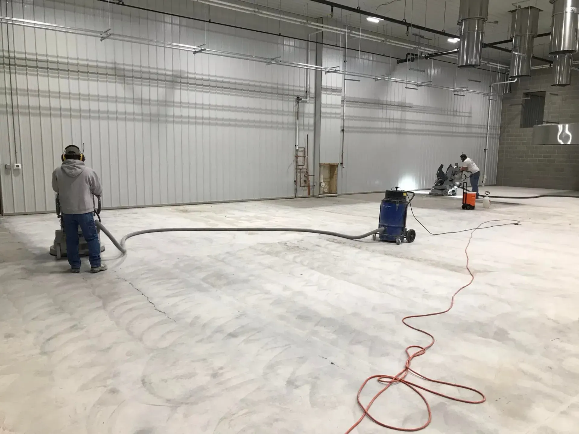 Workers grinding concrete floor in a large, empty warehouse. Dust collection system and power cords visible.