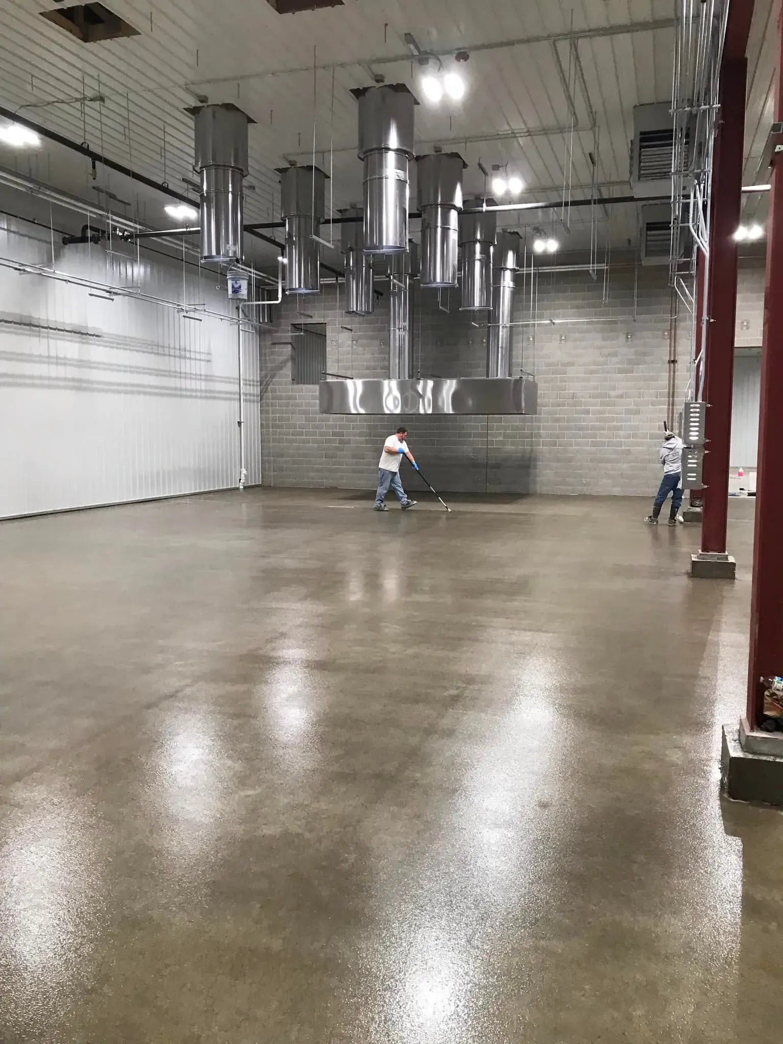 Two people cleaning a large industrial space with shiny, wet floors. Metal vent pipes hang from the ceiling.