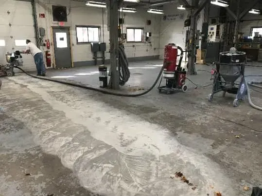 Man vacuuming a dust-covered concrete floor in a workshop. A long, dusty path is visible.