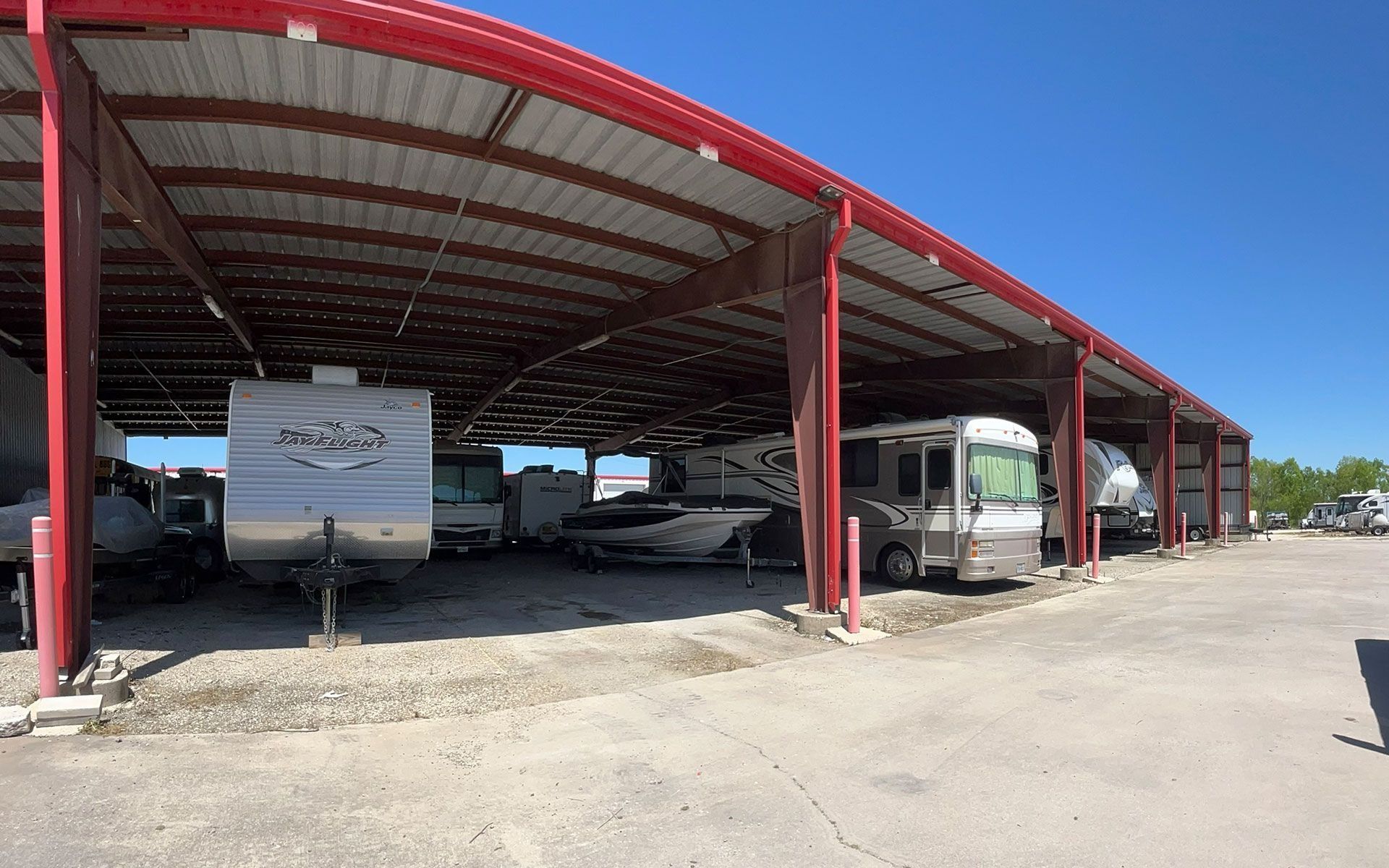 Red-roofed shelter with RVs and boats parked beneath on a gravel lot; sunny day.