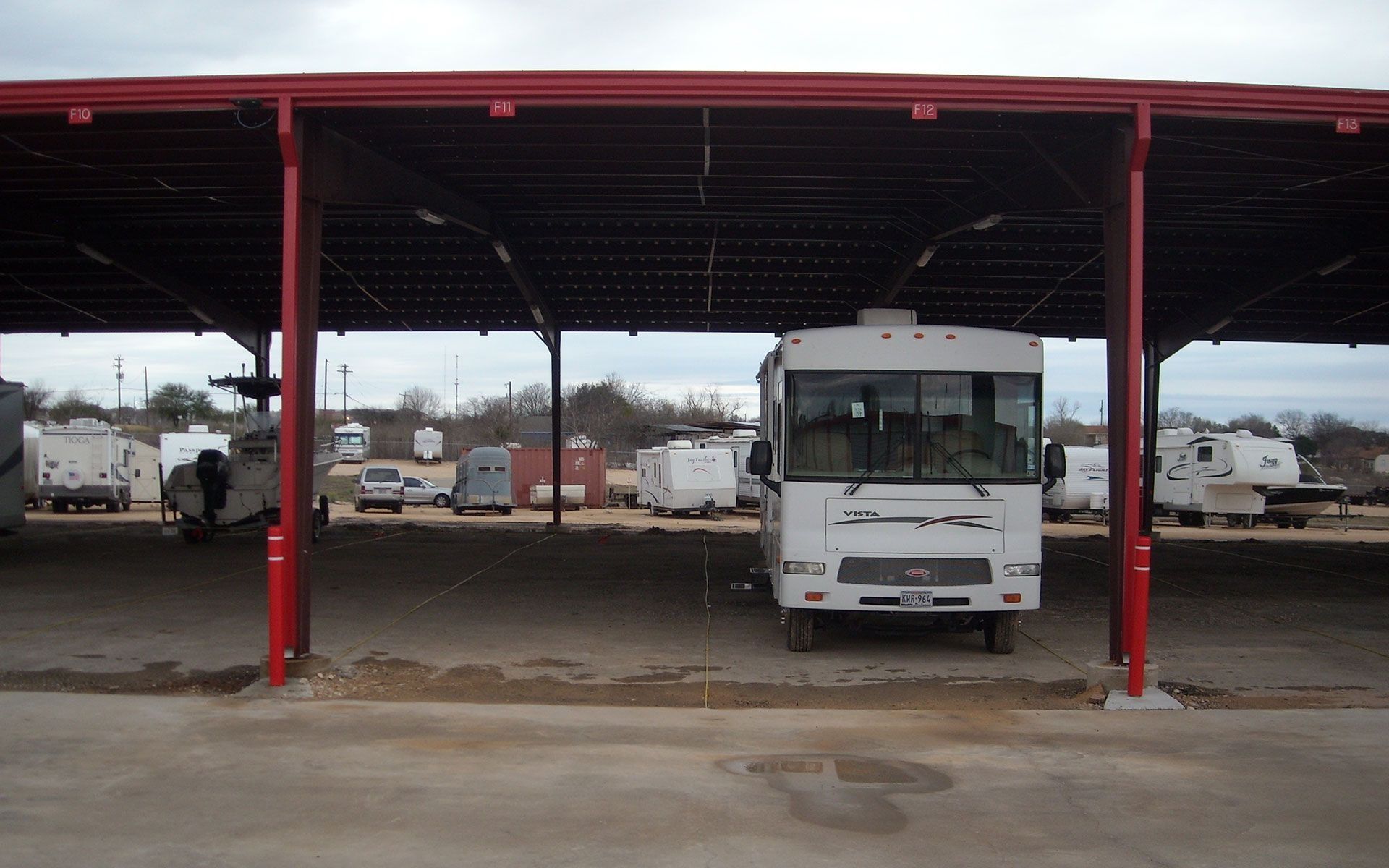 RV parked under a red-roofed carport at a storage facility, other RVs visible in the background.
