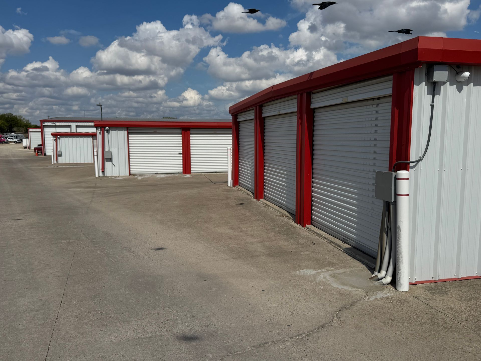 Storage units with red trim and white roll-up doors. Parking lot and blue sky with clouds.