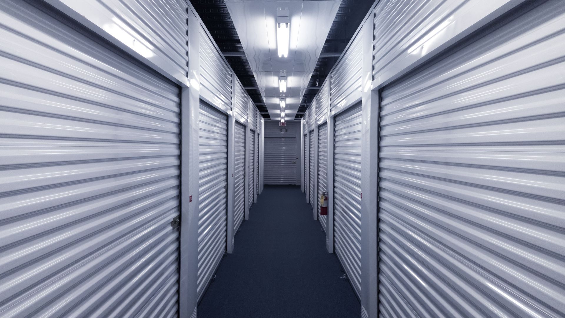 Hallway of storage units with metal doors, white walls, and fluorescent lighting.