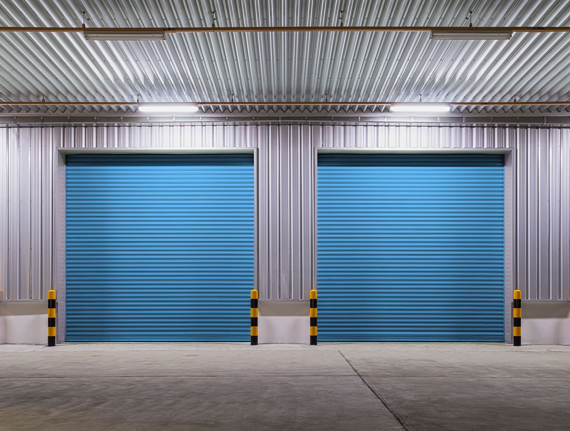 Two blue roller doors in a warehouse, with safety poles and a corrugated metal ceiling.
