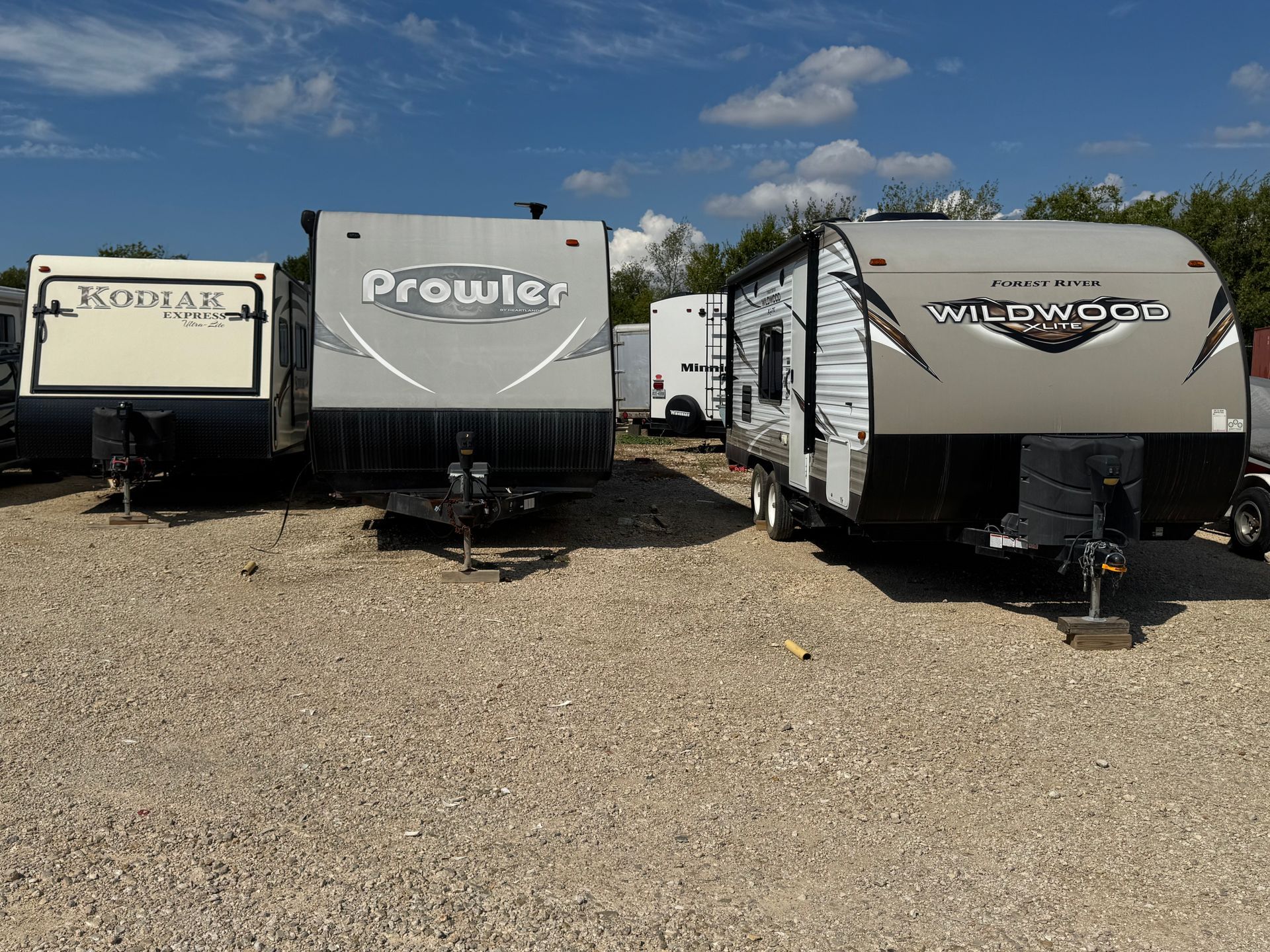 Three RV trailers parked on gravel under a blue sky.