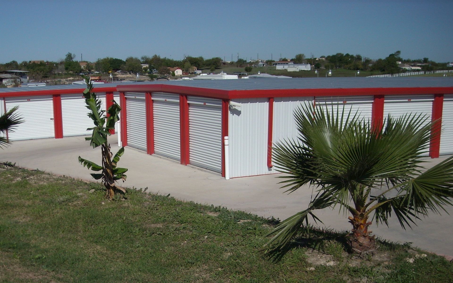 Storage units with red trim and white doors, with palm trees and grass in foreground.
