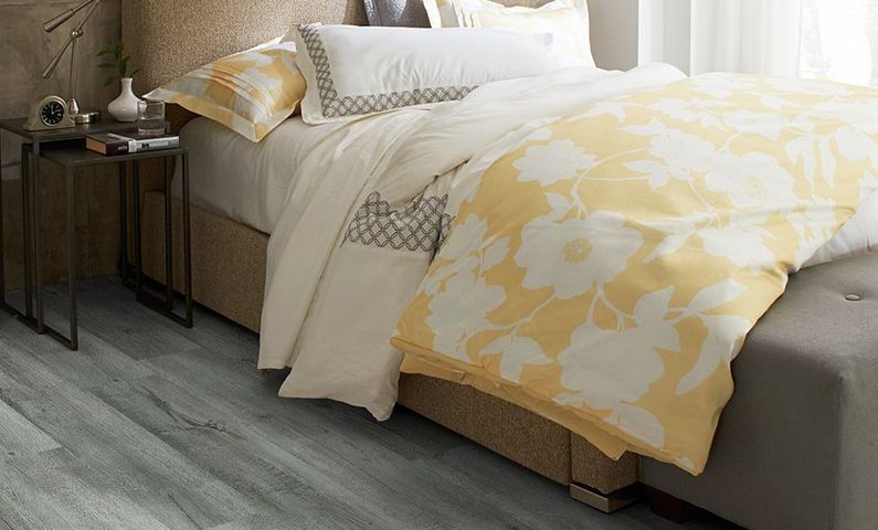 Bedroom with a made bed in white and yellow linens, a side table, and wood-look flooring.