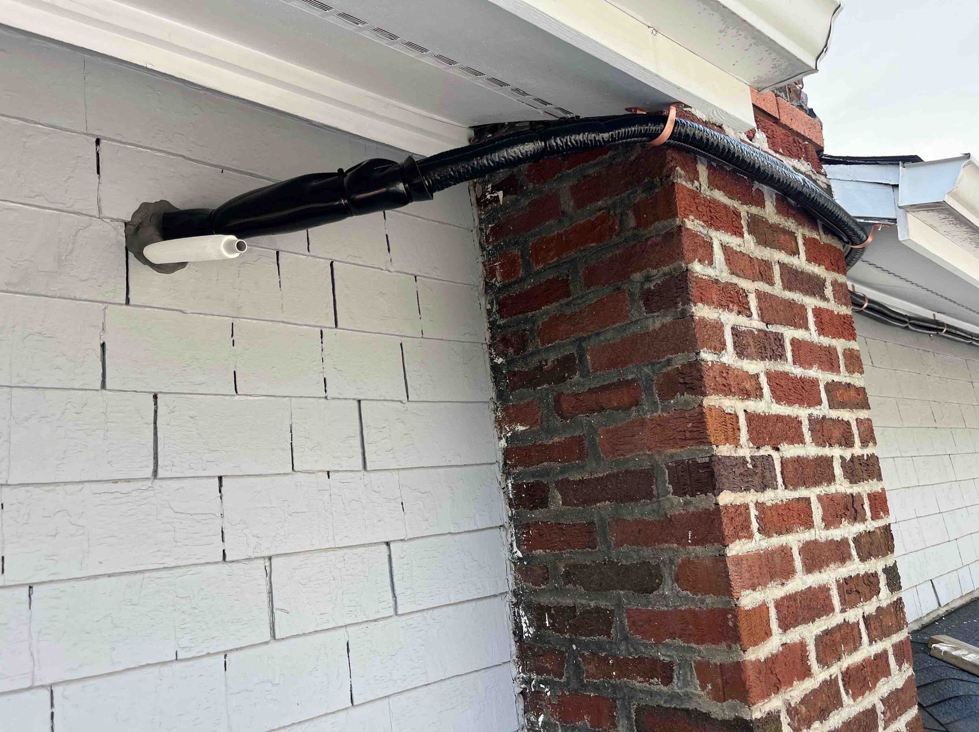 Black tubing exiting a white wall, running alongside a brick chimney.