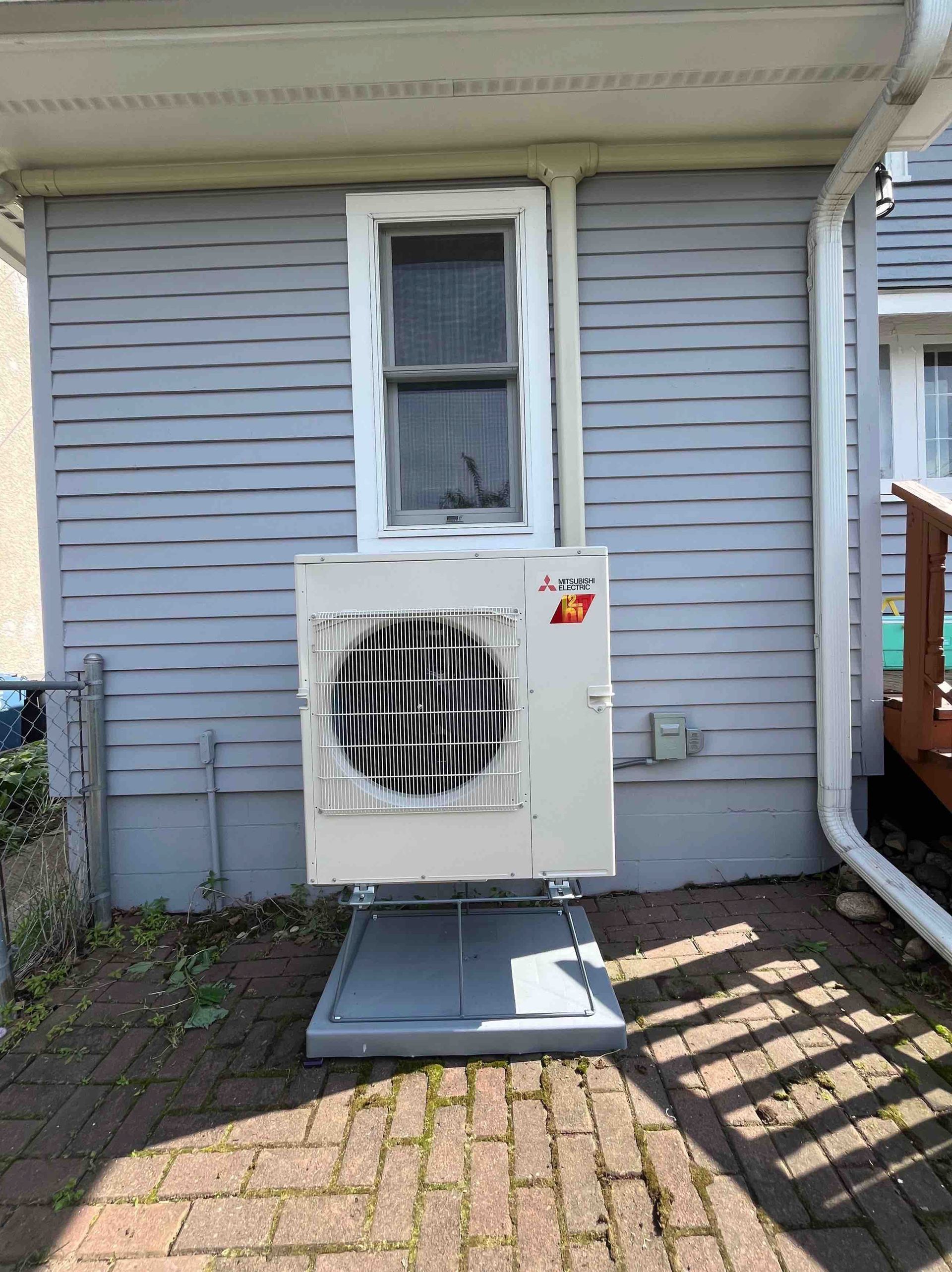 Gray exterior air conditioner unit mounted below a window on a light gray clapboard house.
