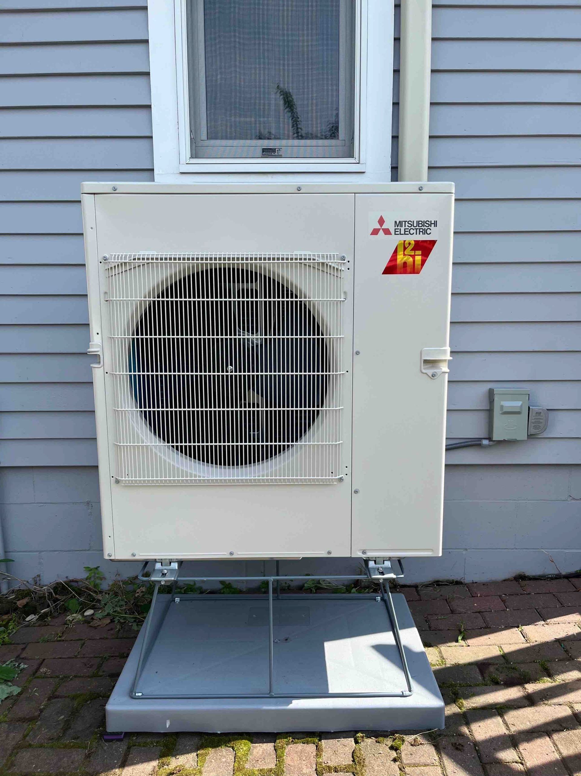 White Mitsubishi heat pump unit mounted on a grey base, next to a window on a blue siding wall.