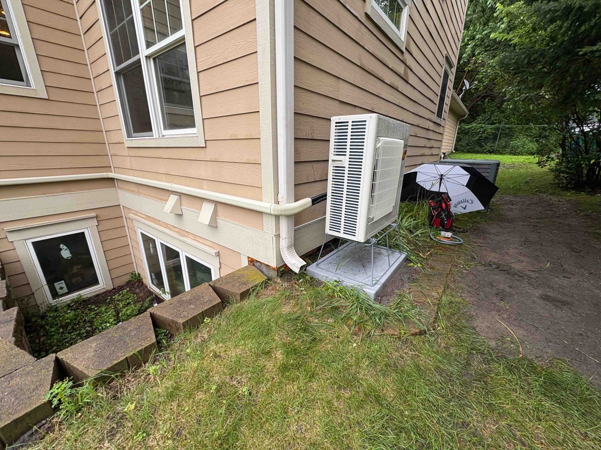 An air conditioner unit installed outside a light tan house with a driveway.
