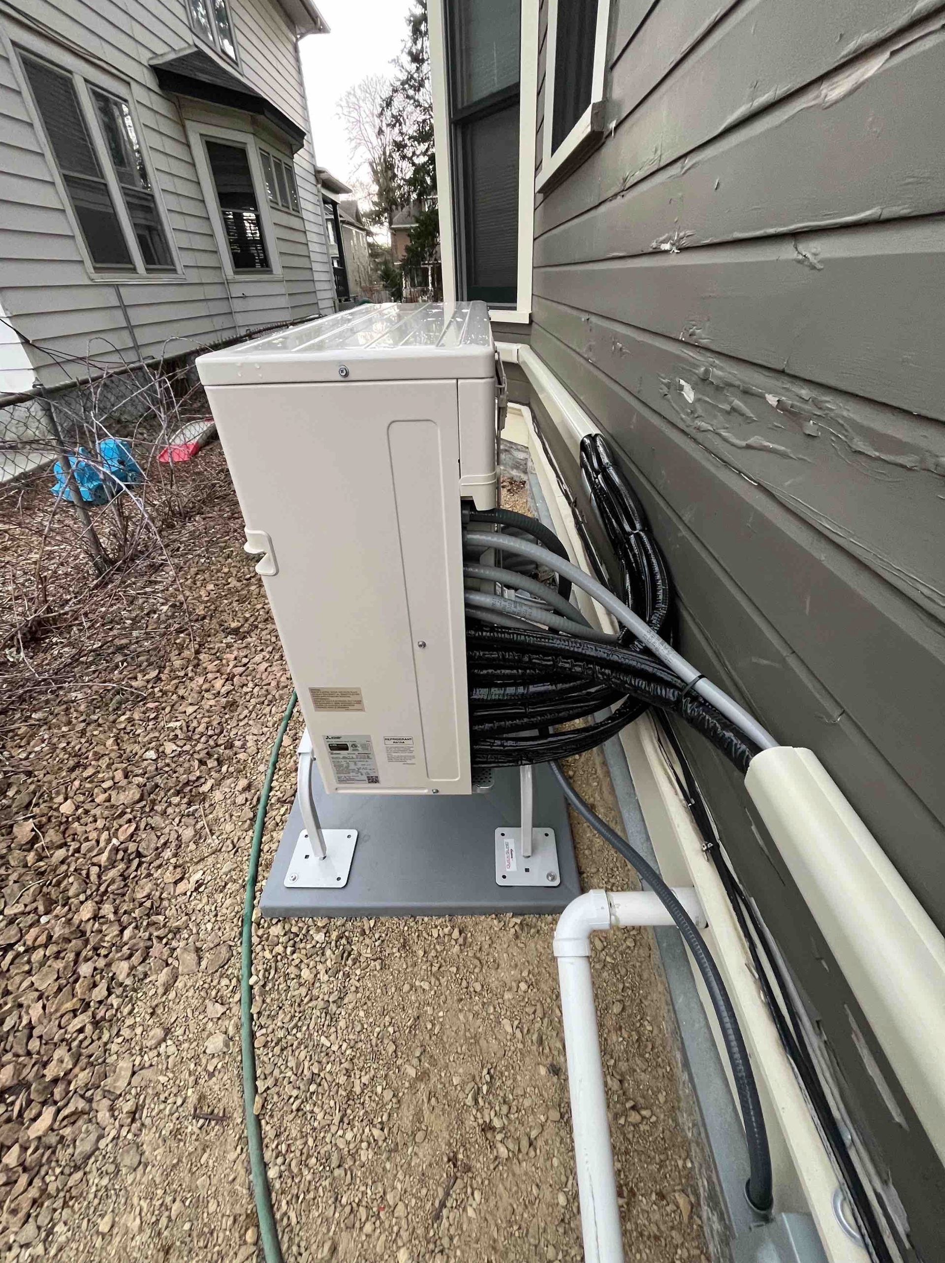 Outdoor HVAC unit mounted on a concrete pad next to a house with exposed wiring and pipes.