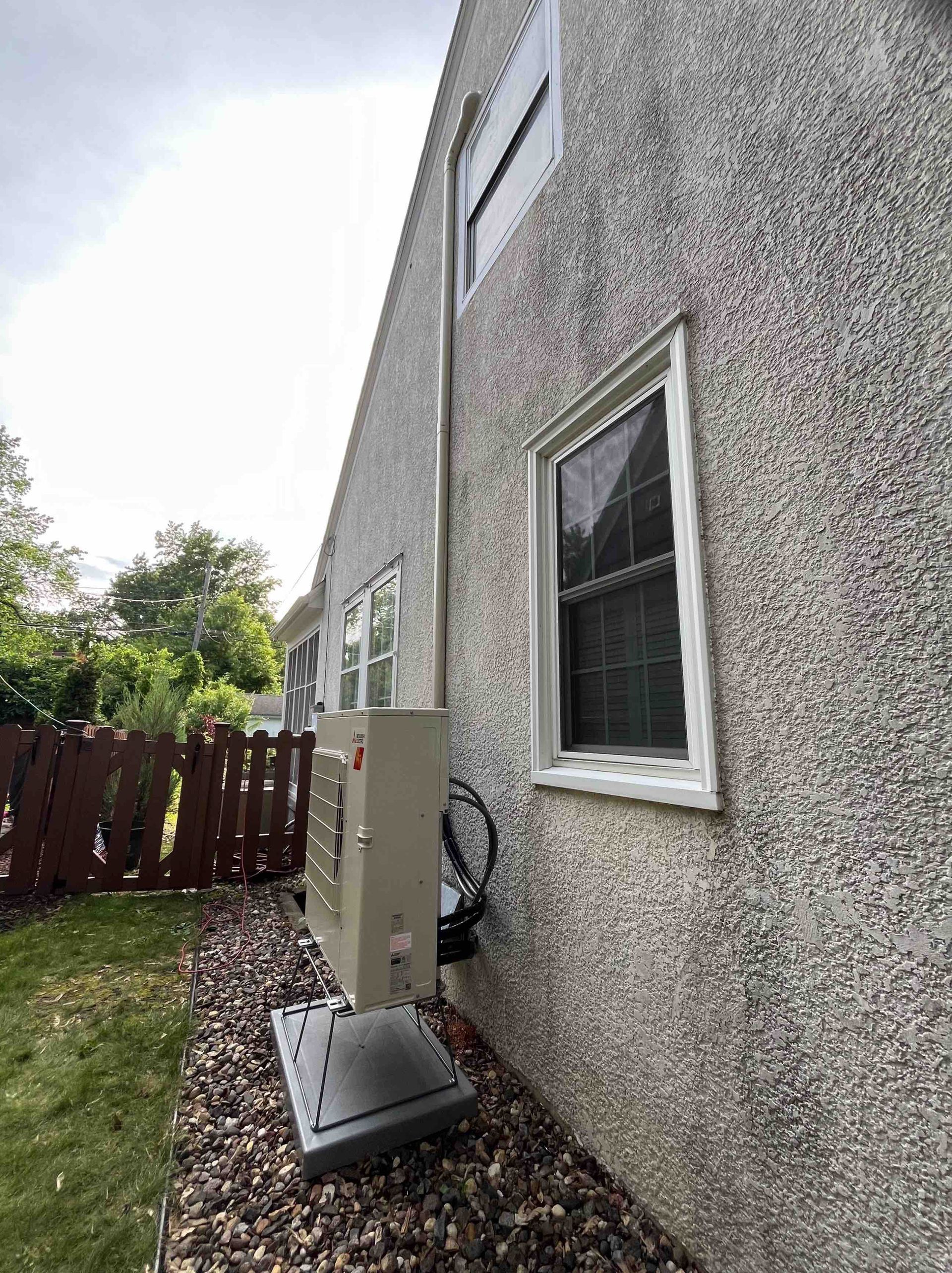 Exterior of a house with an air conditioning unit and exhaust pipe mounted on the stucco wall, next to windows.