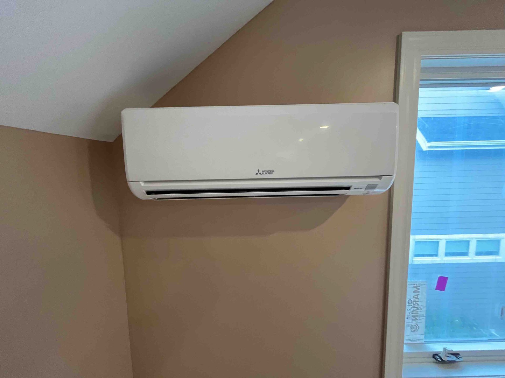 Wall-mounted, white air conditioner on a tan wall in a room with a window.