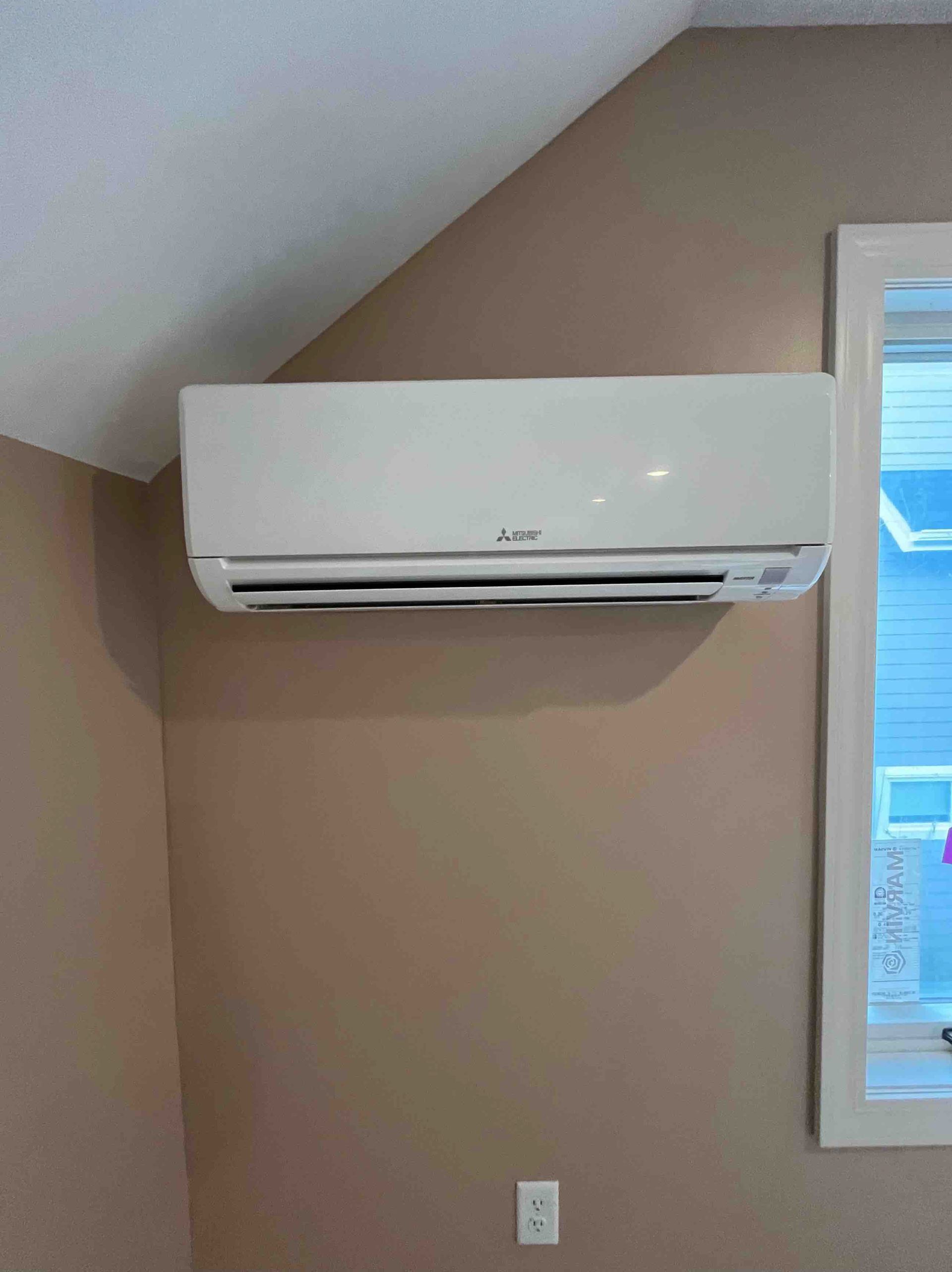 White air conditioner mounted on a tan wall next to a window in a room.